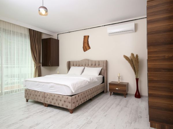 A modern, bright bedroom with a tufted bed, white bedding, a wooden nightstand, a floor vase with dried plants, and a large window with curtains.