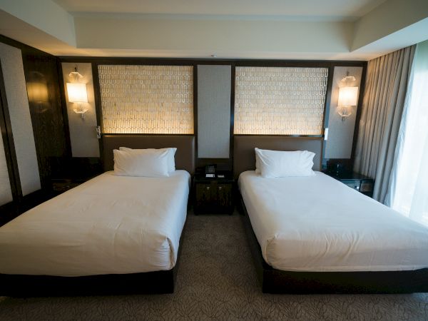 Two twin beds in a modern hotel room with matching headboards, nightstands, and soft wall lighting, plus a window with sheer curtains.