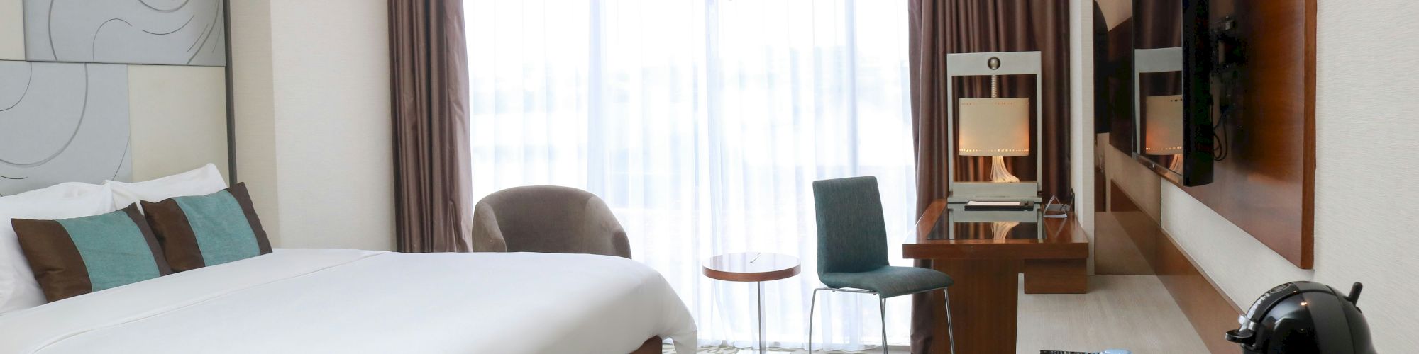 A modern hotel room with a large bed, wall-mounted TV, desk and chair, a seating area near a window, and a neat setup with bottled water and tea/coffee on the table, all in neutral tones.